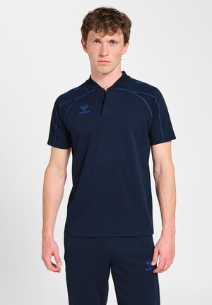 Mand stående iført en marineblå kortærmet sportstrøje og matchende bukser med blå syninger og logoer på bryst og lår.