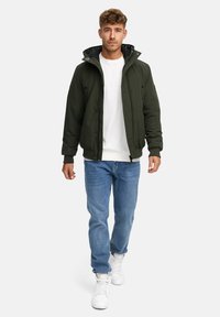 Chaqueta bomber verde con capucha, cierre de cremallera, puños acanalados y una textura suave, combinada con vaqueros azules y zapatillas blancas.