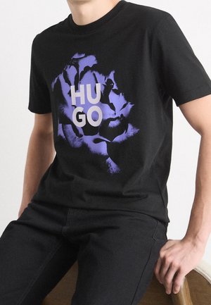 Mies, jolla on musta T-paita, jossa suuri violetti kukka ja valkoinen "HUGO"-teksti, istuu oikea käsi lepäämässä puisella tuolilla.