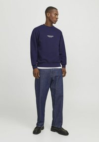 Sudadera marinera de cuello redondo con logotipo blanco, combinada con jeans de mezclilla holgados. El modelo lleva zapatos negros. Se puede ver una camiseta blanca lisa por debajo.