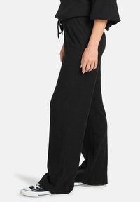 Zwarte wijde broek van ribbelstof met een trekkoord in de taille en zijsplitten, gecombineerd met zwart-witte sneakers.
