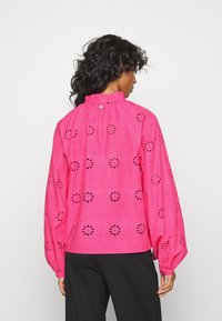 Blusa de linho rosa com padrões florais recortados, mangas longas e bufantes, e uma gola alta com babados, usada com calças pretas.