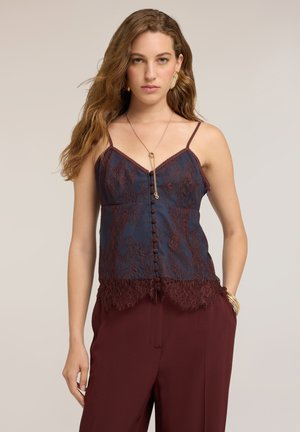 Donna con capelli castani mossi, indossa un camicetta di pizzo blu scuro e pantaloni bordeaux, accessoriata con orecchini, collana e braccialetti d’oro.