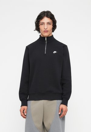 Jeune homme aux cheveux bouclés foncés portant un pull à zip noir Nike et un pantalon en polaire gris clair devant un fond blanc uni.