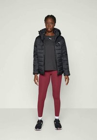 Svart boblejakke med hette, grå treningstopp, burgunder leggings og svarte joggesko. Merkevarelogo som er tydelig på jakken og toppen.