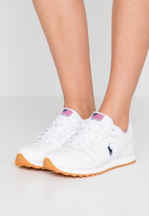 Białe trampki na naturalne stopy, z jasnobrązową podeszwą, z logo gracza polo w kolorze granatowym po bokach i metką z flagą USA na języku.