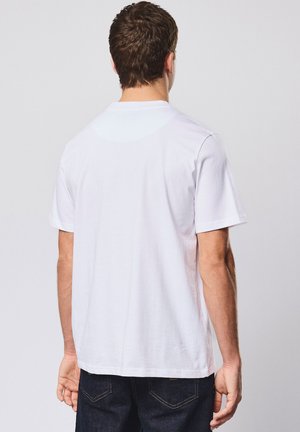 Mann trägt ein einfaches weißes kurzärmeliges T-Shirt und dunkle Jeans, steht mit dem Rücken zur Kamera vor einem schlichten hellen Hintergrund.