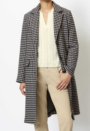 Personne portant un long manteau à carreaux noir et blanc, un pull torsadé crème et un pantalon beige, debout avec une main dans la poche.