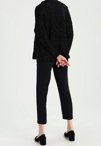 Černý texturovaný kardigan přes upravené černé kalhoty s ohrnutými manžetami a černými botami na blokovém podpatku, pohled zezadu.