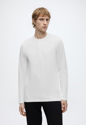 Jonge man met licht haar die een wit Henley-shirt met lange mouwen en zwarte broek draagt tegen een effen lichte achtergrond.