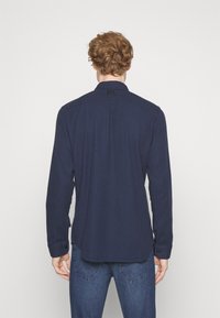 Camicia a maniche lunghe blu scuro con colletto button-down, caratterizzata da una texture liscia e dettaglio sul retro, abbinata a jeans di denim.
