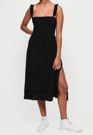 Czarna sukienka do połowy łydki z dopasowanym gorsetem, szerokimi ramiączkami i bocznym rozcięciem. Wykonana z teksturowanej tkaniny z delikatnym wzorem. Noszona z białymi trampkami.