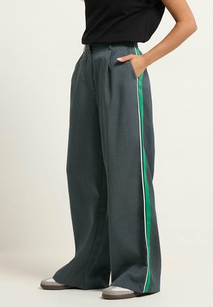 Persona che indossa pantaloni ampi grigio scuro con strisce laterali verdi e bianche, top nero e scarpe da ginnastica grigie, in piedi con una mano in tasca.