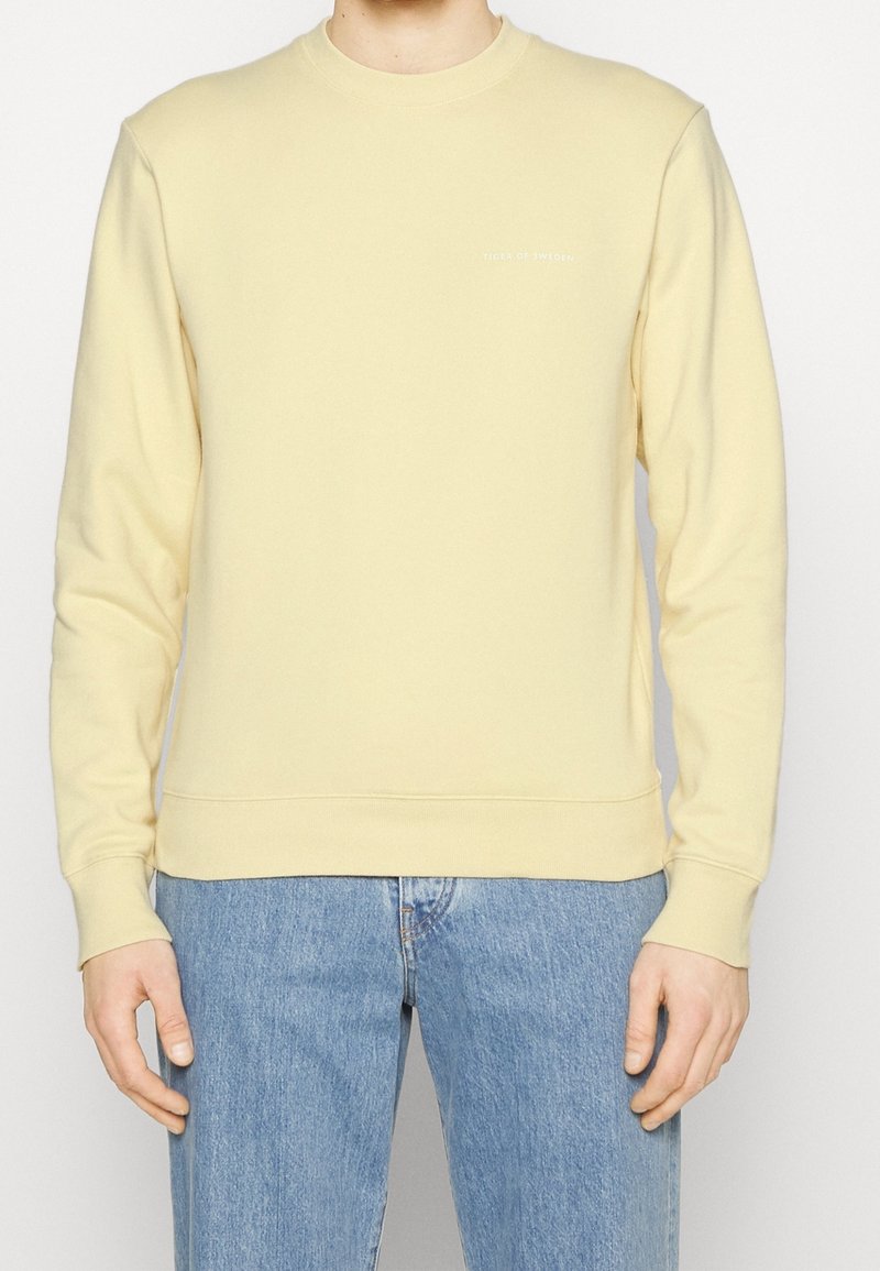 Sudadera de un amarillo claro con escote redondo y mangas largas. Presenta un sutil logo en blanco en el pecho. Tejido suave, puños y dobladillo acanalados.