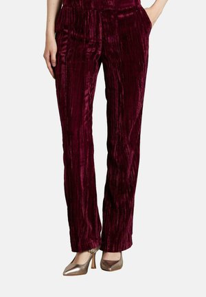 Mujer vistiendo pantalones rectos de terciopelo texturizado rojo profundo y tacones altos metálicos de punta, con las manos en los bolsillos.