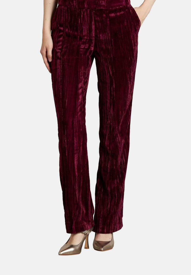 Mujer vistiendo pantalones rectos de terciopelo texturizado rojo profundo y tacones altos metálicos de punta, con las manos en los bolsillos.