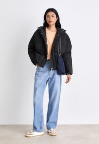 Woman wearing a black hooded jacket, peach shirt, light blue wide-leg jeans, white and beige sneakers, and carrying a navy crossbody bag.