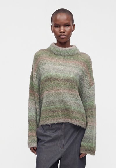 Femme aux cheveux courts portant un pull ample, multicolore vert et gris en matière pelucheuse et un pantalon gris foncé, debout devant un fond uni.