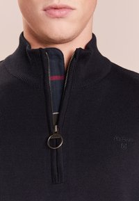 Dunkelblauer gestrickter Viertel-Reißverschluss-Pullover mit hohem Kragen, ausgestattet mit einem Metallreißverschluss und einem karierten Innenfutter mit roten und weißen Akzenten. Barbour-Logo vorhanden.