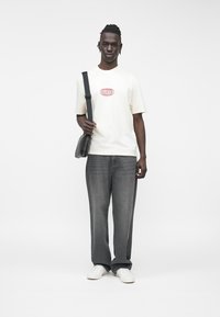 Biała koszulka z krótkim rękawem z czerwonym logo "GUESS", w połączeniu z szarymi jeansami o prostych nogawkach oraz białymi tenisówkami, trzymająca czarną torbę na pasku przez ramię.