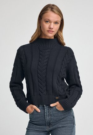 Strikket sweater i mørk marineblå med høj ribbet krave, prydet med kabelstrik mønstre og en tætsiddende ribbet kant. Stylet med denim jeans.