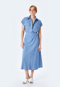 Abito camicia a righe blu con maniche corte e due tasche frontali, vita aderente, gonna fluida, abbinato a mule bianchi con punta aperta.