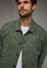 Olivově zelená bunda s texturovaným vzorem, s předním zapínáním na knoflíky, klasickým límcem a dvěma předními kapsami, na bílé košili.