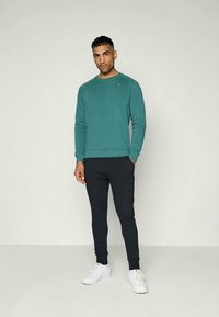 Sudadera turquesa de manga larga y cuello redondo, combinada con joggers negros ajustados y zapatillas blancas. Diseño simple y moderno.