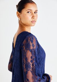 Vestido azul-marinho de renda com padrões florais nas mangas transparentes, corpete ajustado e design com costas em V. Textura suave em toda a peça.