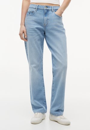 Personne portant un jean droit bleu clair, des baskets blanches et un haut beige, debout avec une main dans la poche.