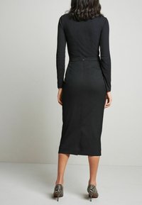 Un haut noir à manches longues associé à une jupe midi noire ajustée, avec une fente au dos et une fermeture éclair discrète. Des talons gris à imprimé serpent complètent le look.