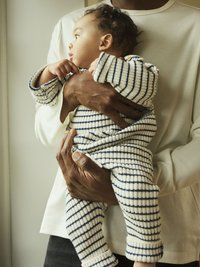 Bawełniany body w kremowym kolorze z granatowymi paskami, z guzikami na ramieniu, trzymane przez osobę w jasnej koszuli.