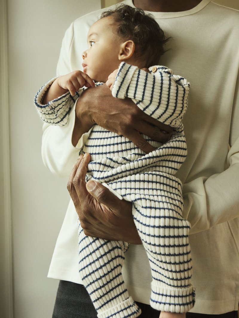 Bawełniany body w kremowym kolorze z granatowymi paskami, z guzikami na ramieniu, trzymane przez osobę w jasnej koszuli.