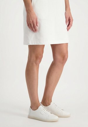 Frau trägt einen weißen Rock und weiße Schnürsneaker, steht vor einem schlichten weißen Hintergrund.