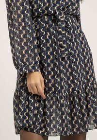 Robe bleu marine avec un motif géométrique beige et marron ; possède des manches longues, une taille ceinturée et un ourlet froncé.