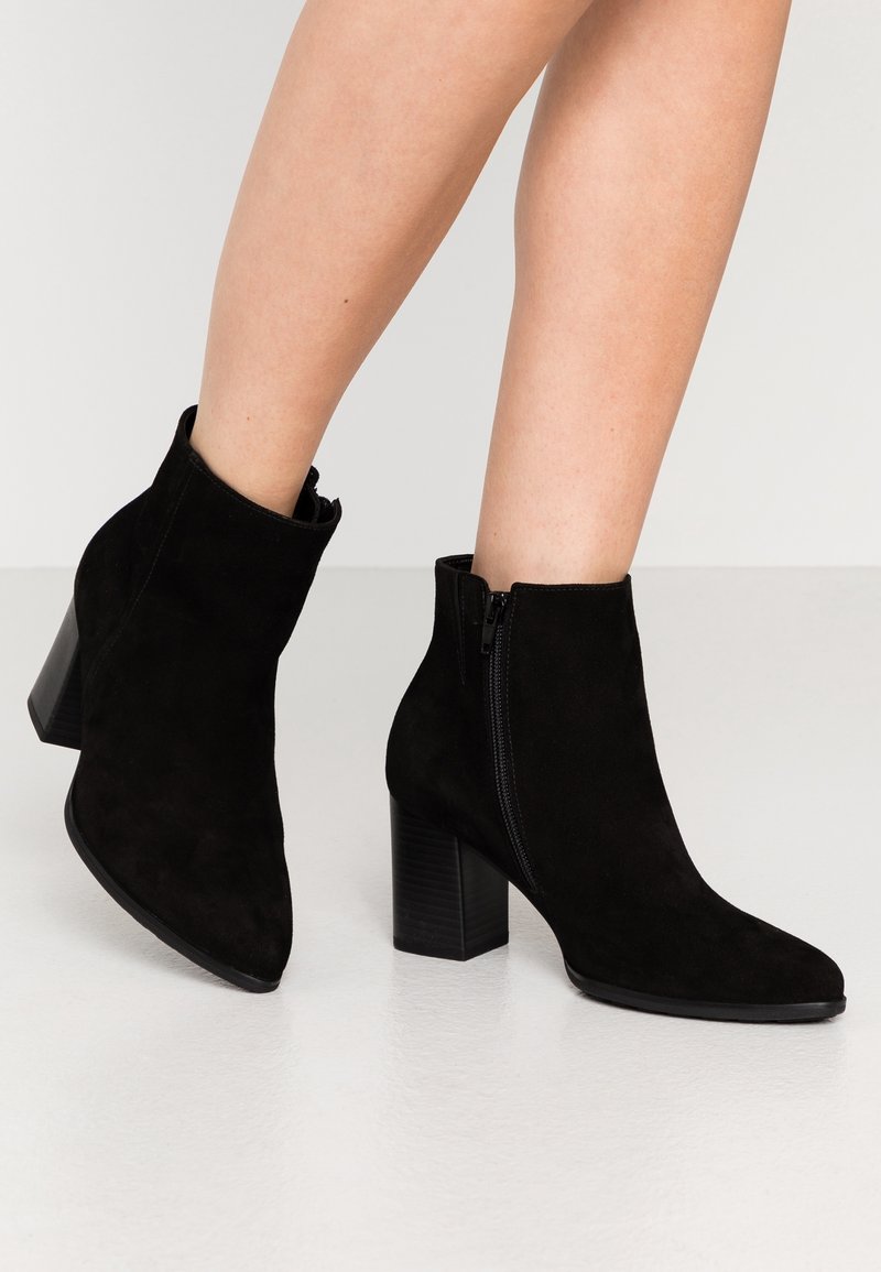 Femme portant des bottines en daim noir à talons carrés et fermetures éclair latérales, debout sur un sol blanc lisse avec un fond neutre.