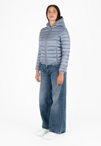 Giacca puffer grigia chiara con zip frontale e cappuccio, caratterizzata da cuciture orizzontali, abbinata a jeans blue larghi e scarpe da ginnastica bianche.