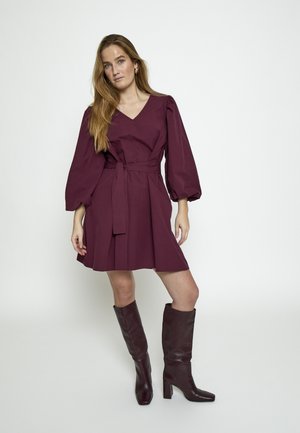 Žena s dlhými blond vlasmi oblečená v burgundských šatách po kolená s nafúknutými rukávmi a k nim ladiacimi vysokými čižmami na päte, stojaca pred bielym pozadím.