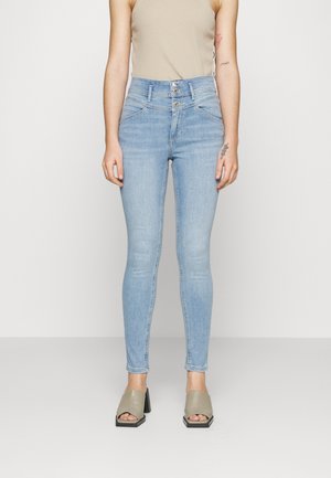 Jeans ajustados de color azul claro con cintura alta, que presentan un cierre de dos botones y detalles de costura sutiles. La modelo lleva una camiseta sin mangas beige y sandalias grises.