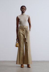 Top bej fără mâneci cu guler înalt, asortat cu pantaloni kaki cu picior larg și curea din material legată. Modelul ține o geantă mică galbenă.