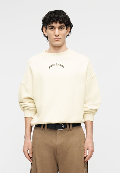 Νεαρός άνδρας με κοντά, σγουρά μαλλιά, φορώντας ανοιχτόχρωμο φούτερ Palm Angels και καφέ παντελόνι, στέκεται μπροστά σε απλό ανοιχτό φόντο.