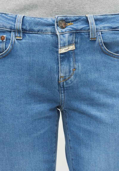 Gros plan sur la taille d'un jean en denim bleu avec un bouton et une petite étiquette en tissu blanc portant l'inscription "CLOD."