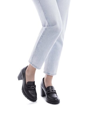 Mocasines de cuero negro con tacón, con un llamativo acento de cadena, parte superior texturizada y una suela gruesa y robusta, combinados con unos jeans de pierna recta de color azul claro.
