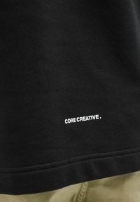 T-shirt in cotone nero con testo stampato bianco "CORE CREATIVE." sull'orlo. Texture morbida e vestibilità classica con dettagli di cucitura.