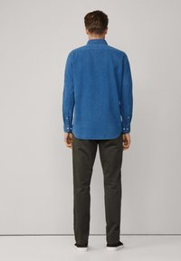 Blauw denim shirt met knoopmanchetten, afgeronde zoom, gedragen over olijfgroene broek en witte sneakers. Aanzicht van achter toont de pasvorm en kleur van het shirt.