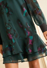 Rochie cu mâneci lungi dintr-un material transparent de culoare teal închis, cu un model de leopard în nuanțe de violet și teal. Dispune de închidere cu nasturi și un tiv cu volane stratificate.