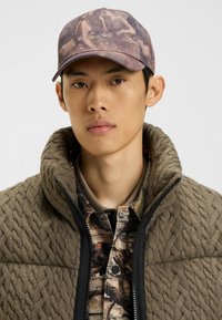 De pet heeft een gestructureerd, abstract camouflagepatroon in gedempte tonen met een verhoogd "HUGO"-logo. Het model draagt een bruine kabelgebreide jas over een gemusterd shirt.