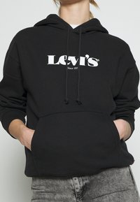 Persona che indossa un hoodie nero di Levi's con tasca frontale e lacci, abbinato a jeans grigi lavati, mani nelle tasche dell'hoodie.