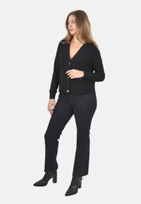 Cardigan nero con scollo a V, maniche lunghe e bottoni dorati, abbinato a jeans scuri e stivali neri con tacco, su uno sfondo bianco.