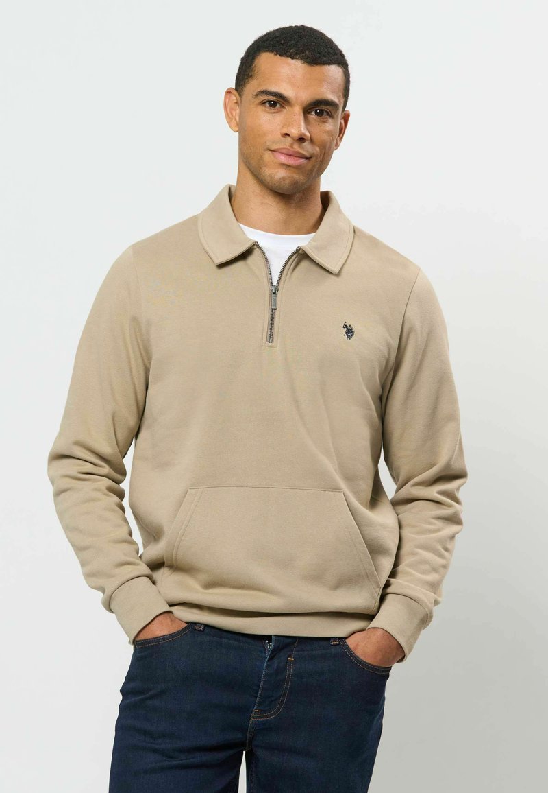 Homme portant un pull beige à fermeture éclair quart de zip avec petit logo et un jean bleu foncé, debout les mains dans les poches devant un fond uni.